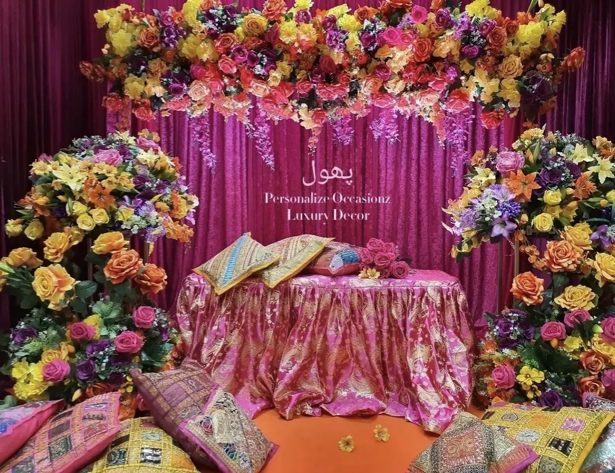 A dholki setup with bright florals, pink drapes and patterned cushions.