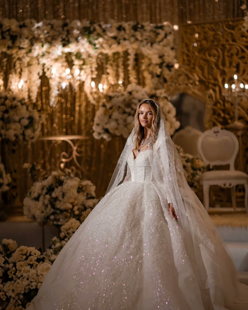 A bride in a sparkling gown before an ivory flower stage.