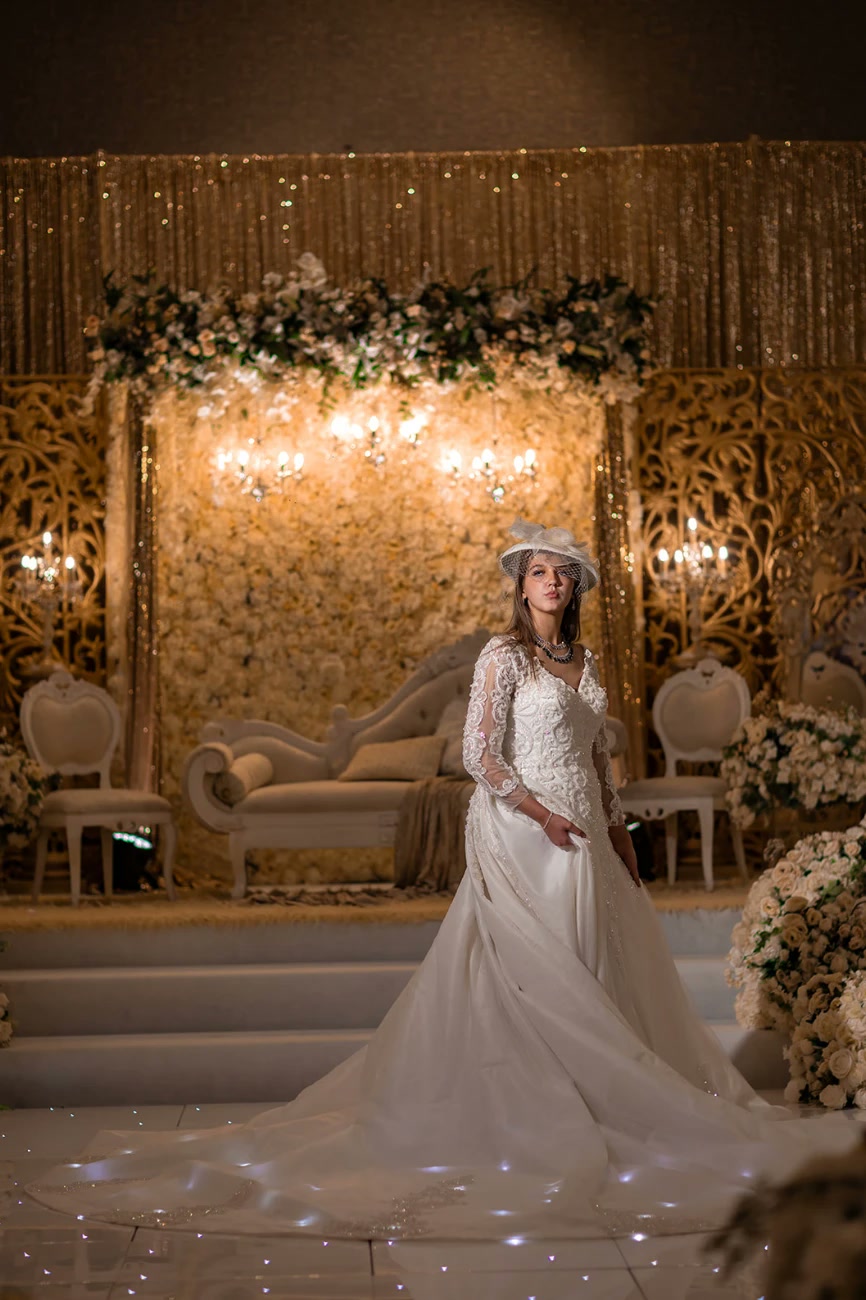 A bride in a lace gown and hat on a white stage with a starlit floor.